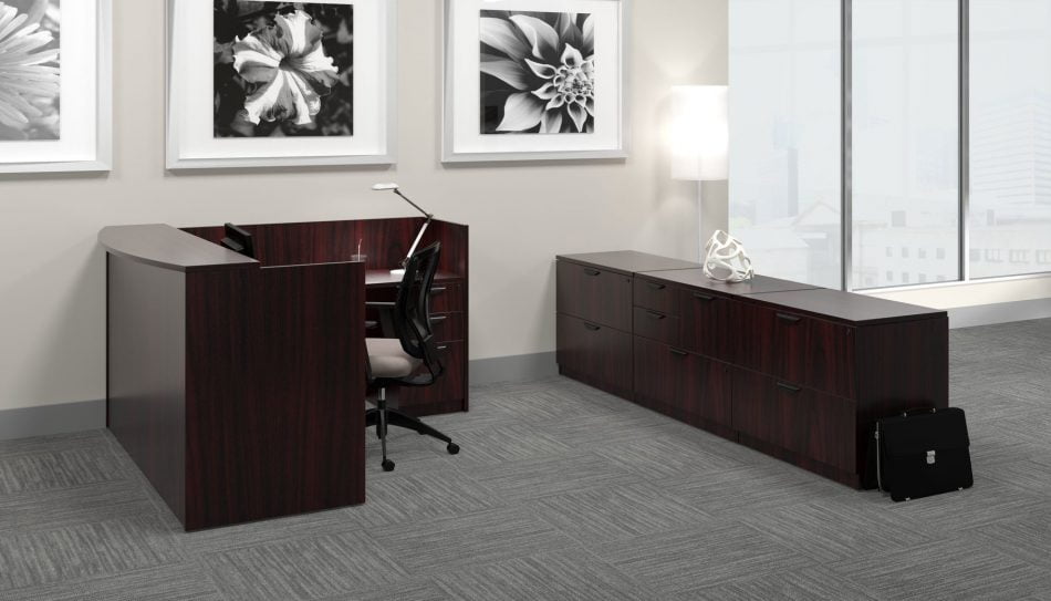Studio photography from the side of an Offices to Go reception desk set. An L- shaped reception desk is against the wall, with a transaction top facing front. A long credenza is placed behind the receptionist chair, with a sculpture placed on top. The furniture is using an American Mahogany finish.