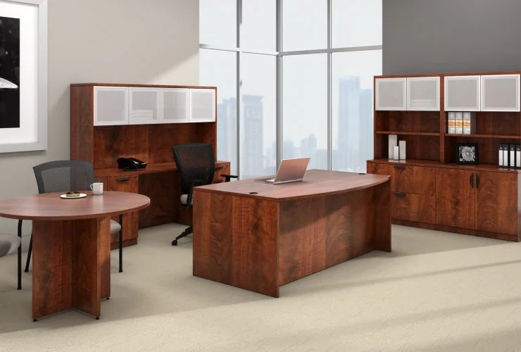 Studio photography of Offices to Go furniture, placed in the corner of a large open office space. A rolling chair is pushed into this space, with a bow front desk. A laptop open and placed on top. To the side is a matching American Dark Cherry laminate round table, with a cup of coffee set on top. Against the two adjoining walls is a mixed storage unit with hutch on top. Each one uses silver framed double doors with frosted acrylic. Through the full height windows is the city below.