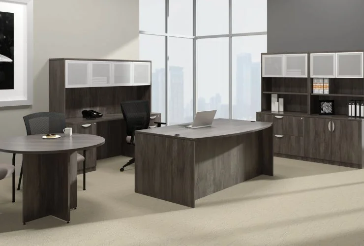 Studio photography of Offices to Go furniture, placed in the corner of a large open office space. A rolling chair is pushed into this space, with a bow front desk. A laptop open and placed on top. To the side is a matching Artisan Grey laminate round table, with a cup of coffee set on top. Against the two adjoining walls is a mixed storage unit with hutch on top. Each one uses silver framed double doors with frosted acrylic. Through the full height windows is the city below.