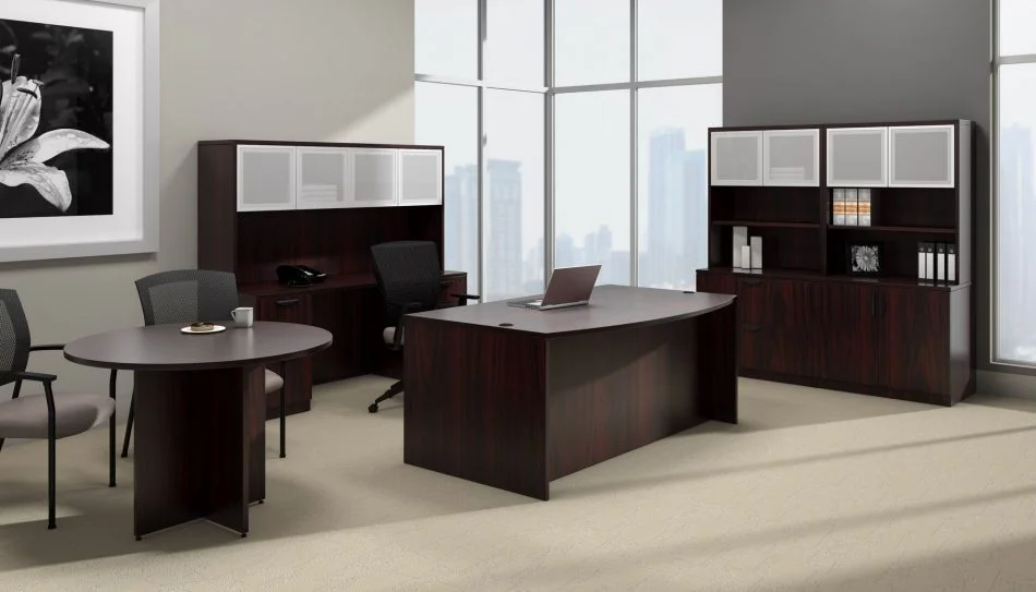 Studio photography of Offices to Go furniture, placed in the corner of a large open office space. A rolling chair is pushed into this space, with a bow front desk. A laptop open and placed on top. To the side is a matching American Mahogany laminate round table, with a cup of coffee set on top. Against the two adjoining walls is a mixed storage unit with hutch on top. Each one uses silver framed double doors with frosted acrylic. Through the full height windows is the city below.