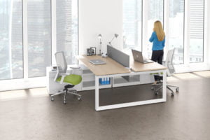 Studio photography of AMQ's Icon Benching. There is a single double run workspace, with a white teak laminate on top. A woman stands and looks out the window, next to a green swivel chair.