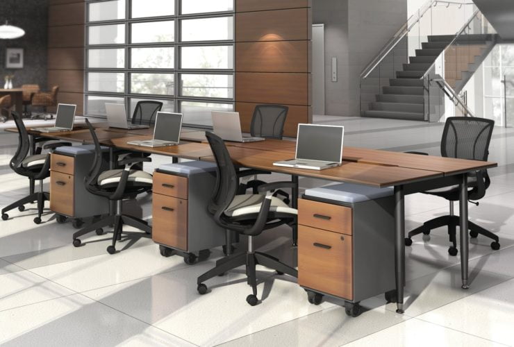 Studio photography of Global's 6-person sidebar benching with a vibrant honey brown finish. Each work station has mobile pedestal drawers and a rolling chair. Beyond, is a flight of stairs and elevator.