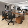Studio photography of Offices to Go's laminate height adjustable workstations. A group of three stations are set together, each with an LED desk lamp and computer. A set of 2 drawers is wheeled in each space, with storage for supplies. Sunlight comes in from windows towards the background. A pair of large black and white paintings hang on the wall off to the right.