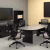 Studio photography of Offices to Go furniture in a conference area. A racetrack shaped table is in the center, with 6 high-back chairs placed around it. On the far wall, a presentation board has one of its double doors open, revealing a white board underneath. At the left is a matching mixed storage unit, with a tall book case at each side, and a flat screen TV directly above. The furniture uses an American Espresso finish.