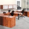 Studio photography of Offices to Go furniture, consisting of two L shape desks arranged from front to back, with a D-island just in between. There are two chairs pulled up to these L-shape workstations, a set of three overhead compartments mounted on the wall above. The furniture and shelving uses an American Dark Cherry finish.