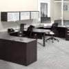 Studio photography of Offices to Go furniture, consisting of two L shape desks arranged from front to back, with a D-island just in between. There are two chairs pulled up to these L-shape workstations, a set of three overhead compartments mounted on the wall above. The furniture and shelving uses an American Espresso finish.