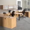 Studio photography of Offices to Go furniture, consisting of two L shape desks arranged from front to back, with a D-island just in between. There are two chairs pulled up to these L-shape workstations, a set of three overhead compartments mounted on the wall above. The furniture and shelving uses an Autumn Walnut finish.