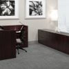Studio photography from the side of an Offices to Go reception desk set. An L- shaped reception desk is against the wall, with a transaction top facing front. A long credenza is placed behind the receptionist chair, with a sculpture placed on top. The furniture is using an American Mahogany finish.